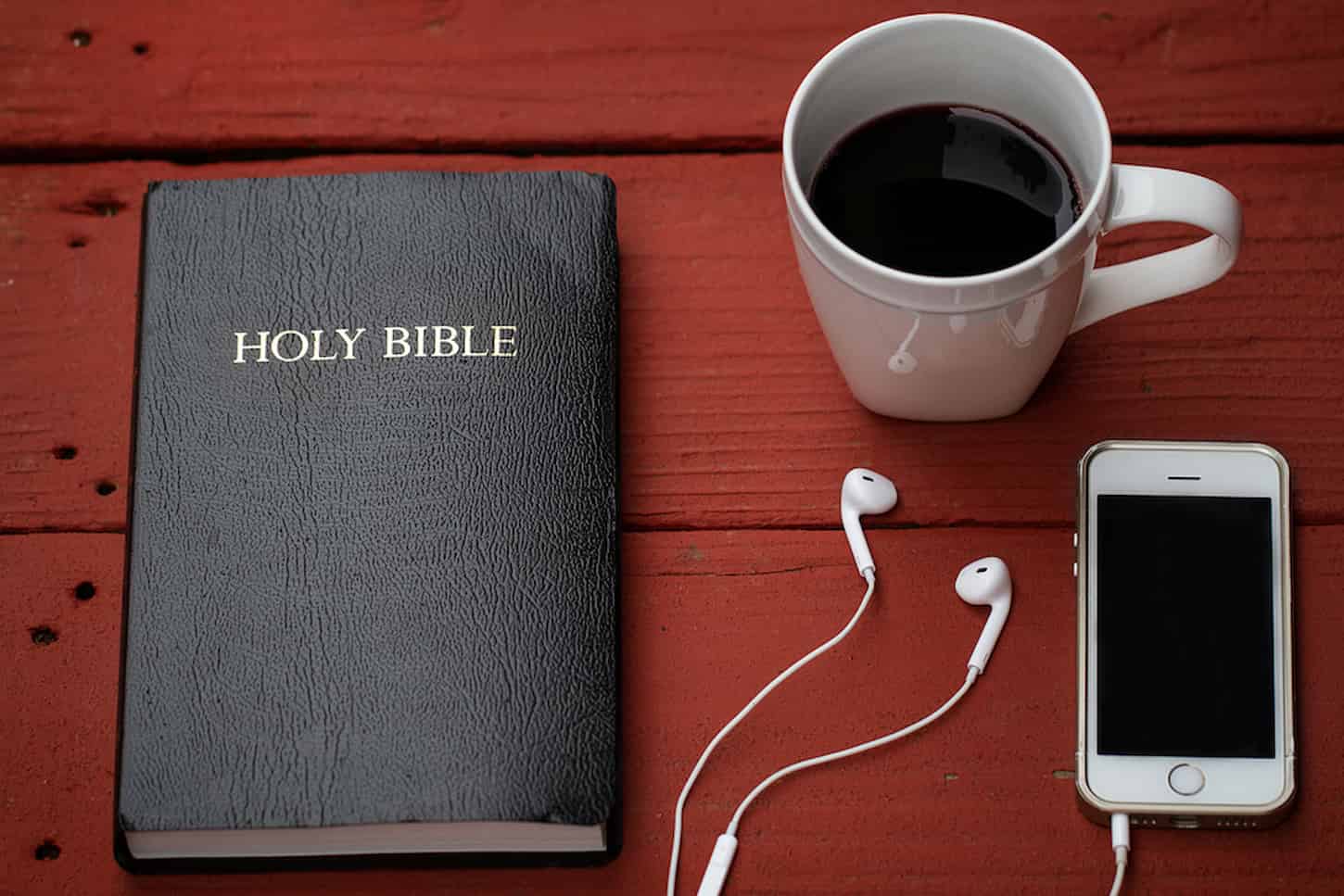 An Image of a bible, coffee in a white mug, and a cellphone with a headset on top of a wooden table.