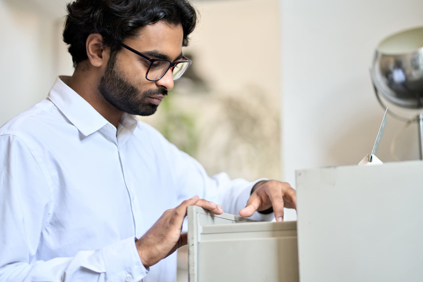 An image of an Indian businessman searching corporate documents in an archive drawer. Files reference directory administrator accountant looking for papers, doing paperwork, organizing folders accounting storage.